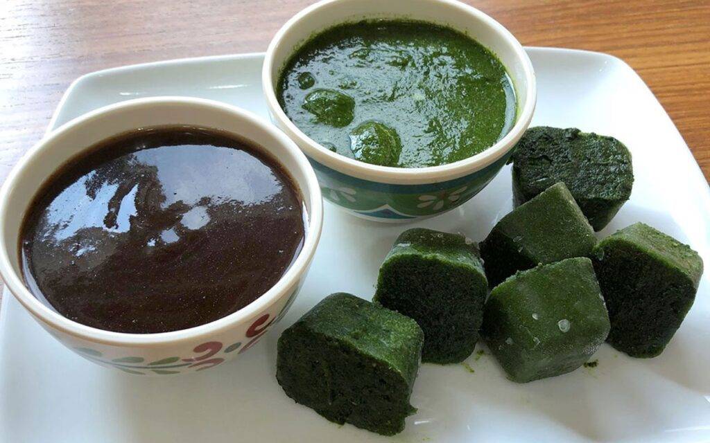 Bowls of tamarind and mint chutneys served with cubes of frozen mint chutney on a white plate.