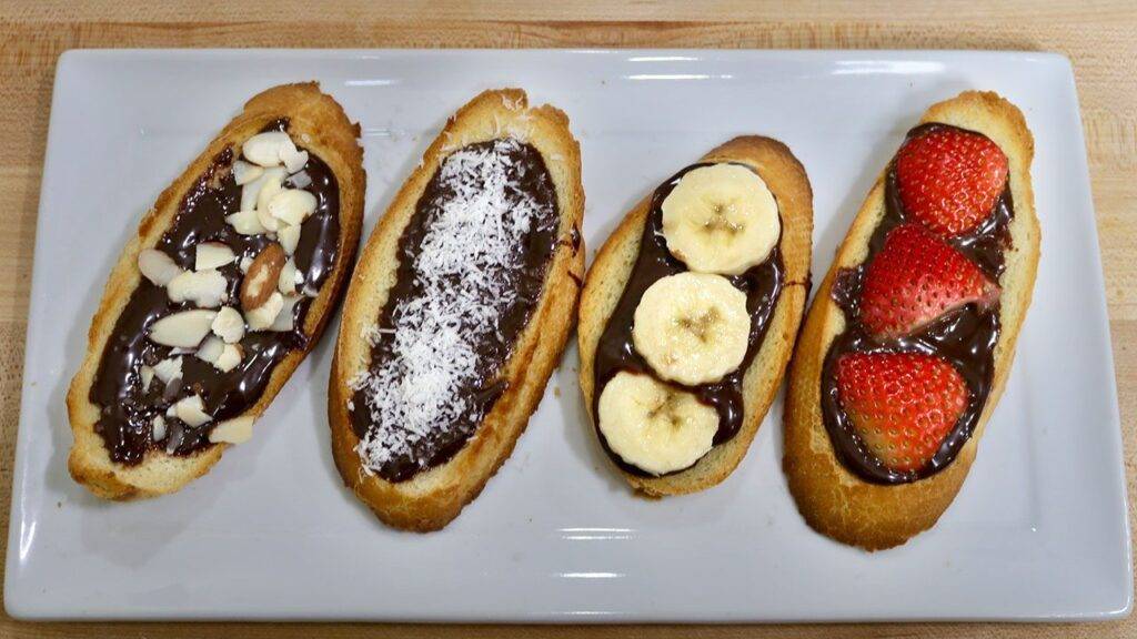 Four slices of toasted bread topped with chocolate ganache and various toppings: sliced almonds, shredded coconut, banana slices, and strawberry halves.