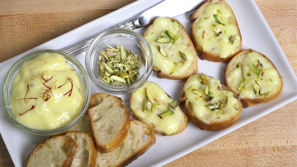 Slices of toasted bread topped with custard and garnished with sliced pistachios, served with extra custard and a bowl of pistachios on the side.