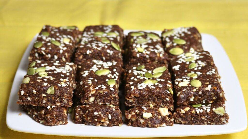 A platter of homemade Dates and Nuts Energy Bars, topped with sesame seeds and pumpkin seeds, arranged neatly on a white plate with a yellow background.