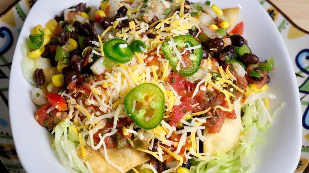 A vibrant plate of Mexican Bhel, topped with black beans, corn, diced vegetables, shredded cheese, and sliced jalapenos.
