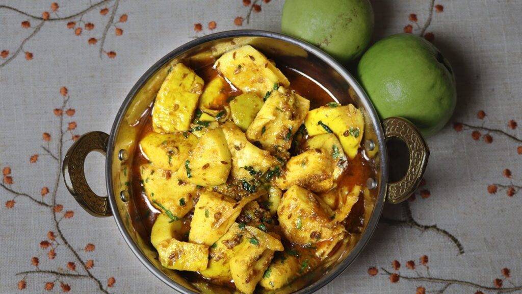 A bowl of vibrant yellow Sweet and Sour Guava Curry, garnished with fresh herbs, with whole guavas placed beside the bowl on a patterned tablecloth.