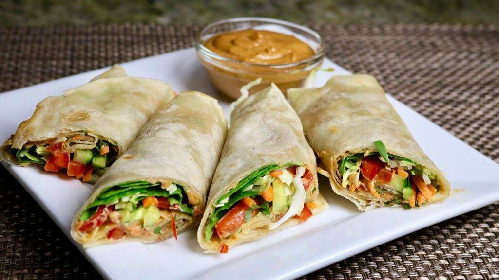Four vegetable kathi rolls filled with fresh veggies, served with a side of dipping sauce.
